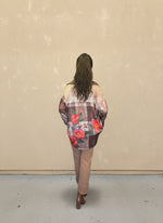Model facing away wearing a brown plaid floral button-up shirt with red rose prints, paired with tan pants and brown ankle boots, standing against a beige wall.