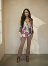 Model posing in a beige and brown plaid floral shirt with red rose design and leopard trim, tucked into tan pants and paired with brown boots in natural light by a window.
