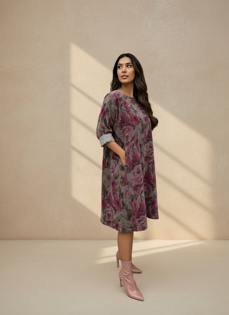 Woman wearing a knee-length loose floral dress in shades of mauve and rose, styled with rolled-up sleeves and pink heeled ankle boots, posing sideways in soft natural light.