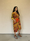Woman wearing a vibrant yellow and red floral dress with cuffed sleeves, styled with brown ankle boots and an orange fuzzy crossbody bag, posing sideways against a beige wall.
