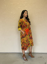 Woman wearing a vibrant yellow and red floral dress with cuffed sleeves, styled with brown ankle boots and an orange fuzzy crossbody bag, posing sideways against a beige wall.