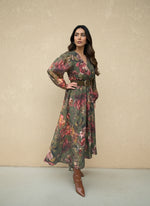 Woman wearing a green floral wrap maxi dress with red and gold floral prints, styled with a wide brown belt and brown ankle boots.