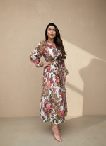Woman in a white floral V-neck maxi dress with puff sleeves and pink floral patterns, styled with blush pink heels, standing against a neutral wall.