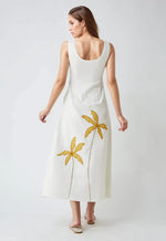 Model wearing sleeveless white cotton dress with mustard yellow palm tree embroidery on the back, standing against a light grey background.