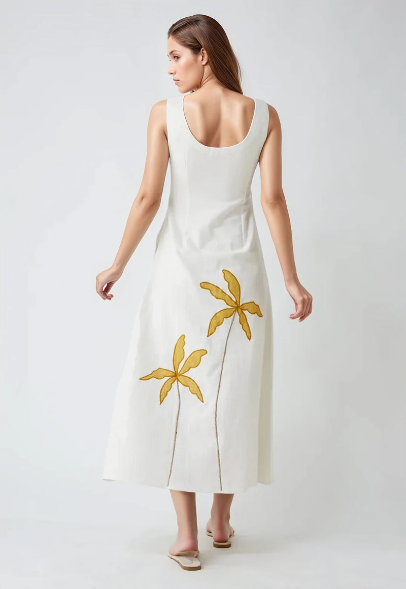 Model wearing sleeveless white cotton dress with mustard yellow palm tree embroidery on the back, standing against a light grey background.