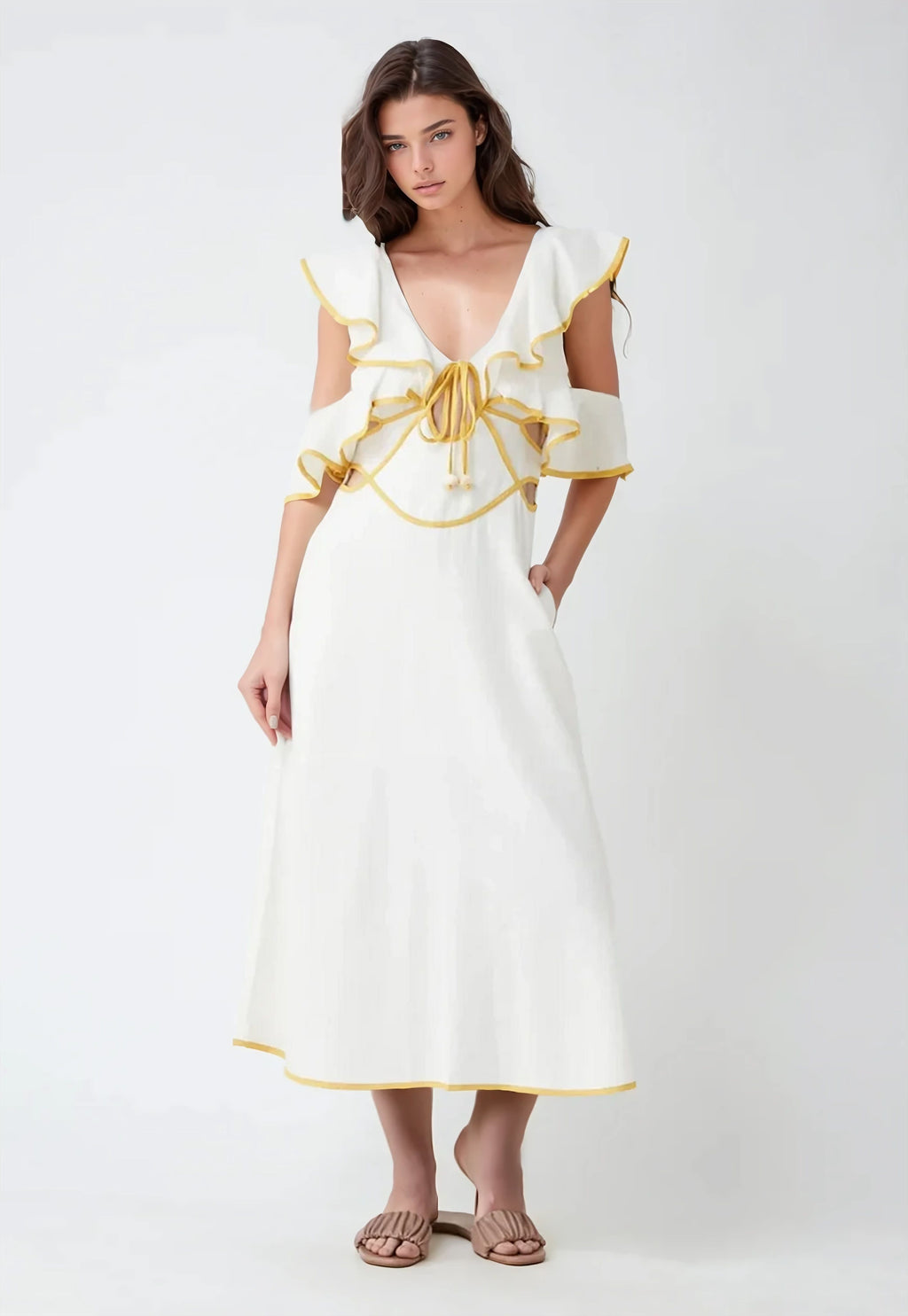 A woman wearing a white midi dress with mustard yellow trim and ruffled sleeves. The dress features a deep V-neckline, open shoulder cutouts, and a tie-front detail with tassels. She stands facing forward with one hand in her pocket.