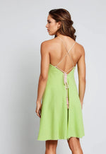 Back view of a woman wearing a lime green cotton mini dress with thin crisscross straps and a tie-back with tassel detail. The dress has a flared shape and raw-edge hem.