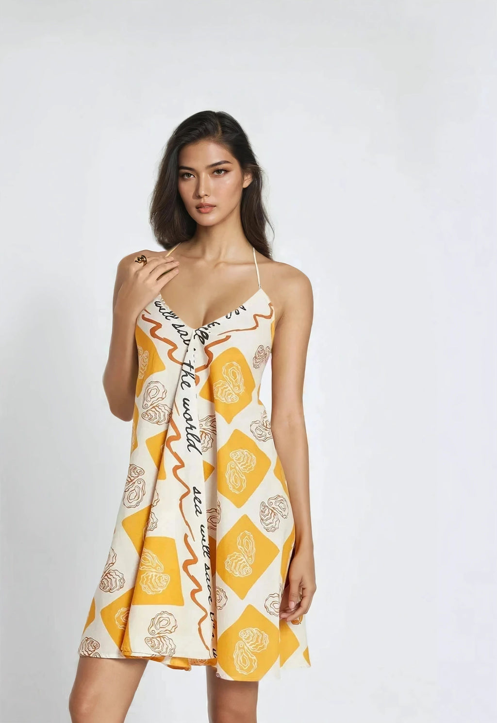 Woman wearing a sleeveless halter dress with a cream and mustard yellow diamond pattern, featuring abstract oyster prints and a script design down the front. The dress has a relaxed A-line silhouette and falls above the knees.