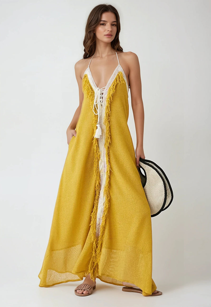 Woman wearing a mustard yellow halter maxi dress with white lace-up front and fringe trim, holding a wide-brim hat.