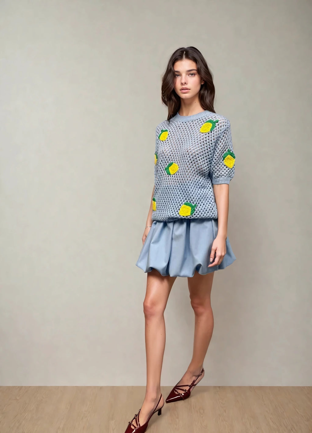 Model wearing a light blue open-knit short sleeve sweater with bright yellow lemon appliqués, paired with a soft blue pleated mini skirt