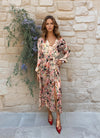 Model wearing a silky blush pink floral midi dress with long sleeves, a V-neckline, and a waist tie belt, standing in front of a stone wall.