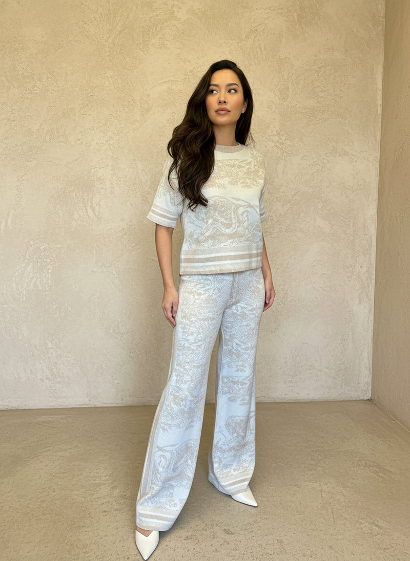 Model wearing a cream and beige jacquard knit two-piece set with subtle baroque print, standing against a textured beige wall.