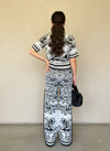 Rear view of a woman in a black and white toile print knit top and pants, her hair styled in a ponytail, holding a black handbag.