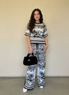 Woman posing in a black and white jacquard knit set with toile-style pattern, black handbag, and white heels against a neutral wall.
