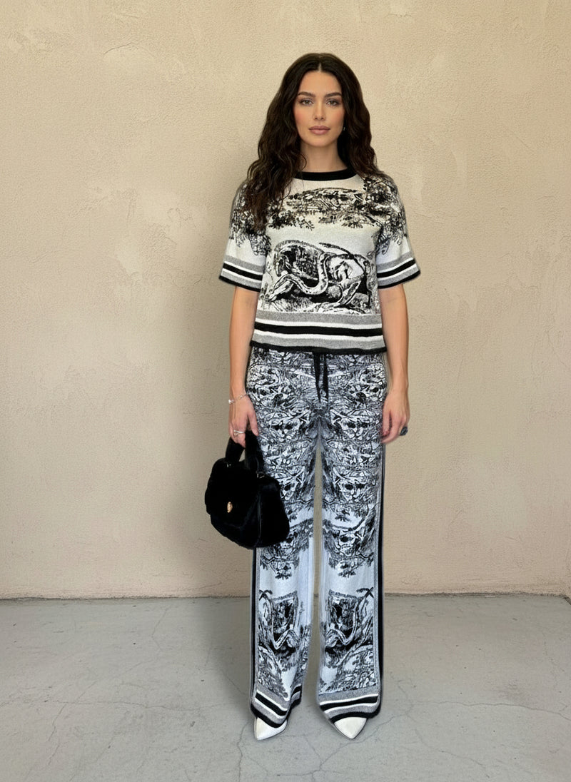 Front view of a woman wearing a black and white printed knit matching set, accessorized with a black faux fur handbag and white pointed-toe heels.