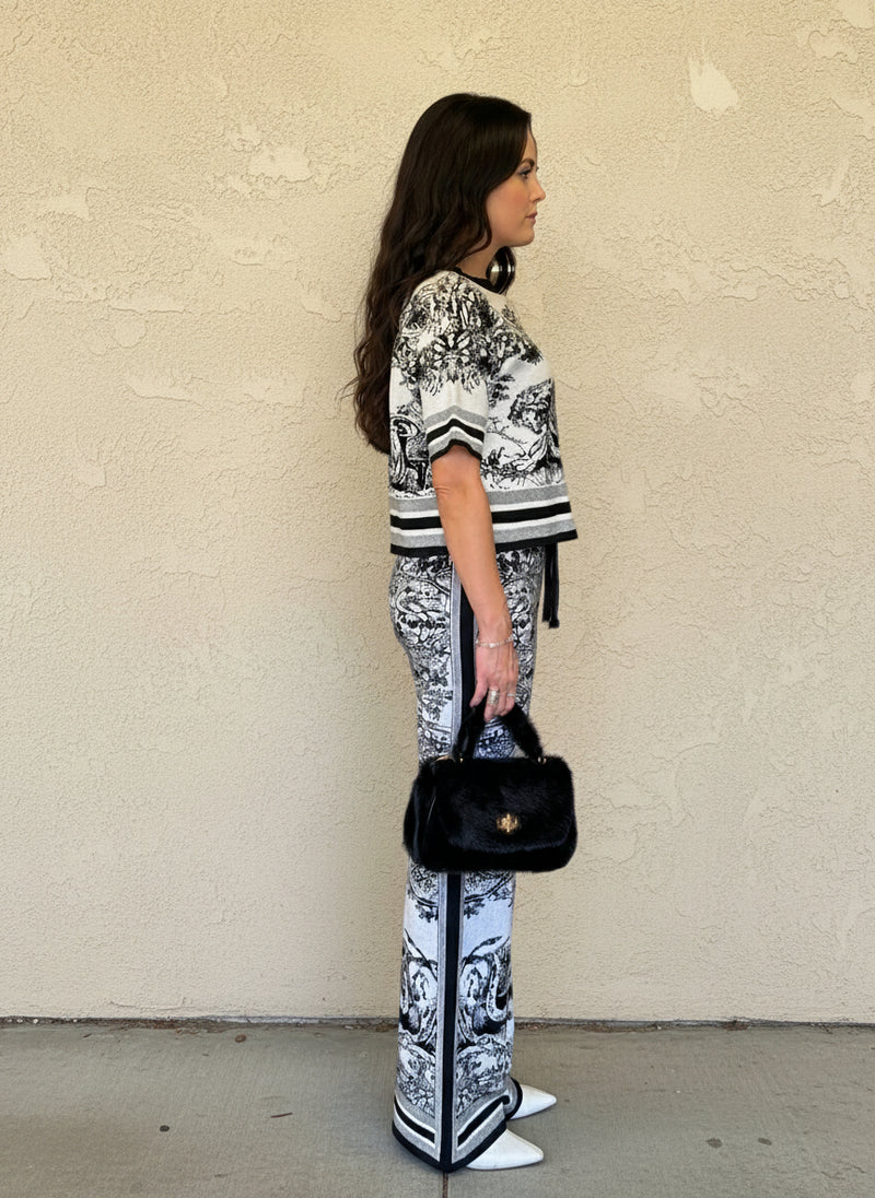 Side view of a woman dressed in a monochrome toile knit two-piece set, accessorized with a black faux fur handbag and white pointed heels.
