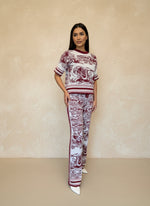 Woman in a burgundy and white toile print knit co-ord set featuring a short-sleeve top and straight-leg pants, posing indoors against a beige wall.
