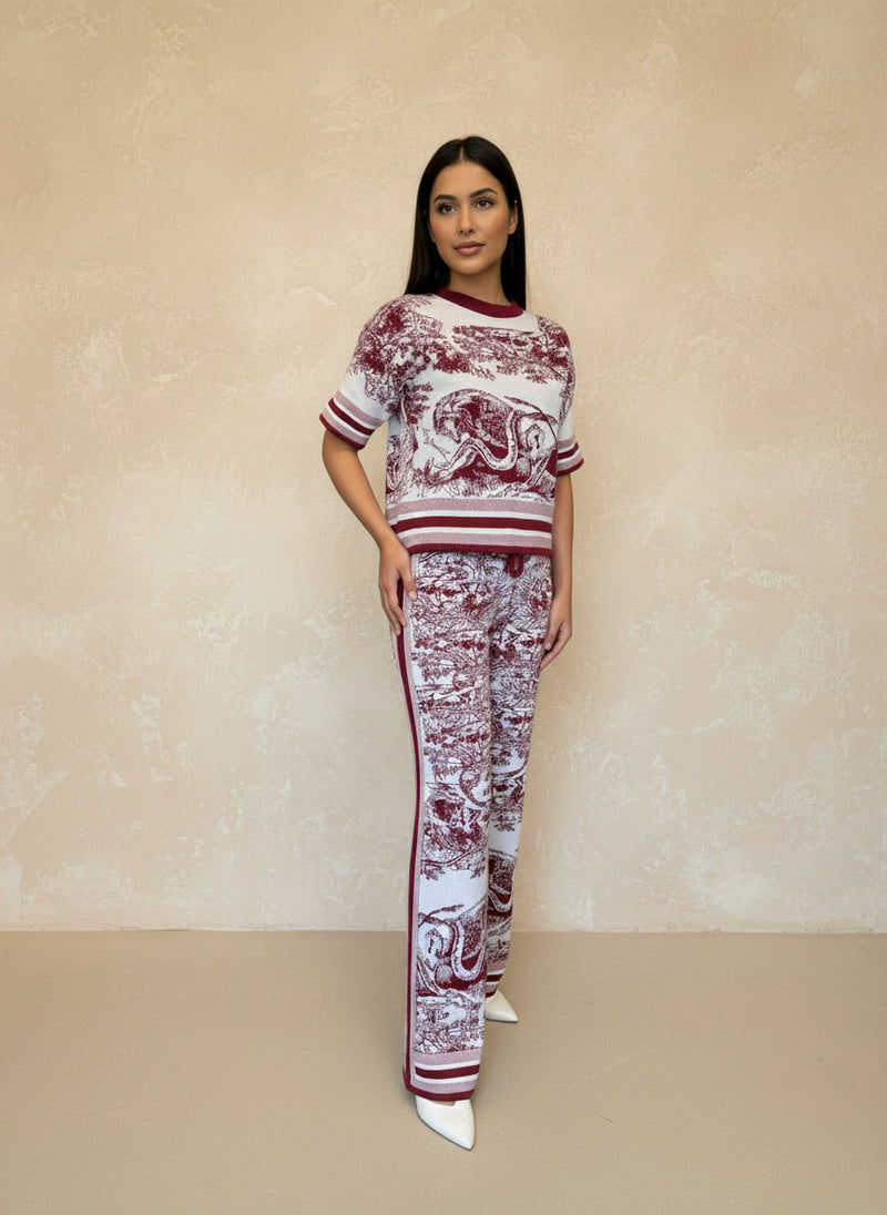Woman in a burgundy and white toile print knit co-ord set featuring a short-sleeve top and straight-leg pants, posing indoors against a beige wall.