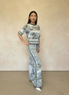 Model posing in a sage green and white printed knit two-piece set with short sleeves and flared pants, styled with white heels.