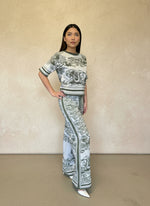 Model posing in a sage green and white printed knit two-piece set with short sleeves and flared pants, styled with white heels.