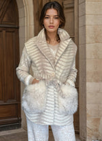 Woman wearing an elegant beige and ivory belted jacket with fur pockets, standing near a stone arch doorway.