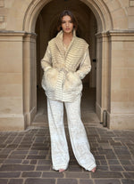 Full-body view of woman in cream patterned pants and beige fur-trimmed belted jacket, posing in an architectural stone archway.