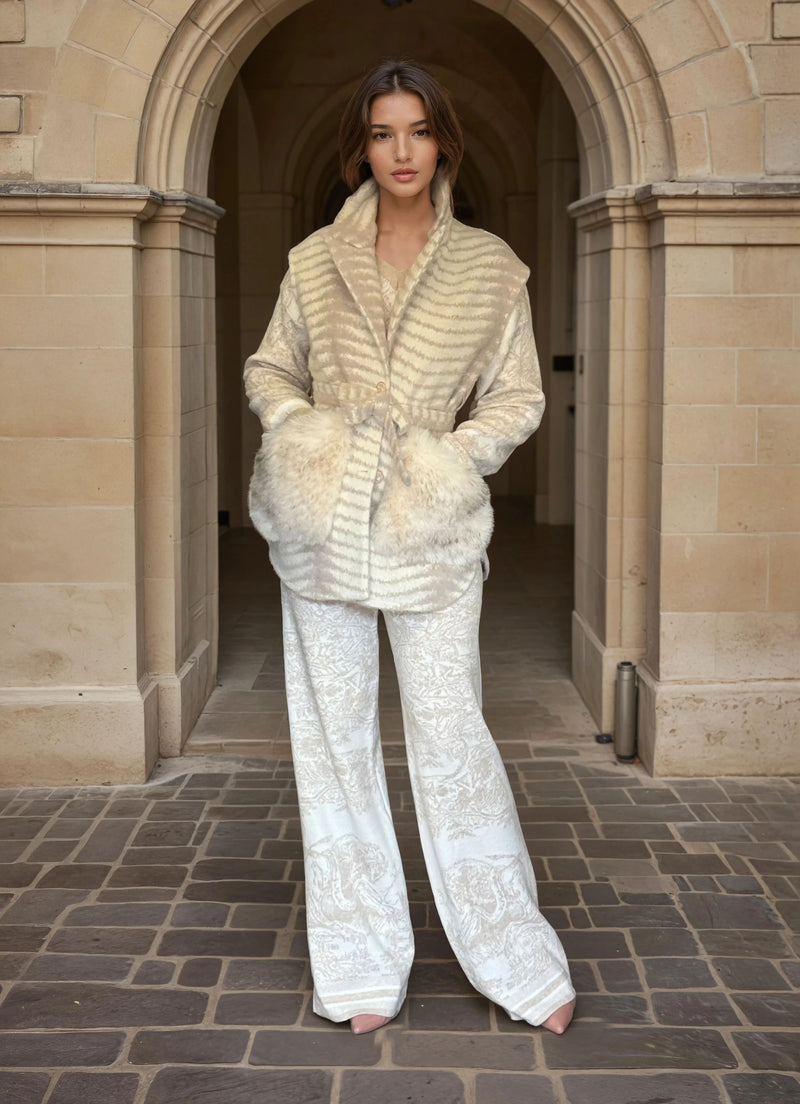 Full-body view of woman in cream patterned pants and beige fur-trimmed belted jacket, posing in an architectural stone archway.