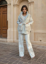 Model in a cream-striped belted jacket with white fur pockets and patterned wide-leg trousers, standing outside a classical building.