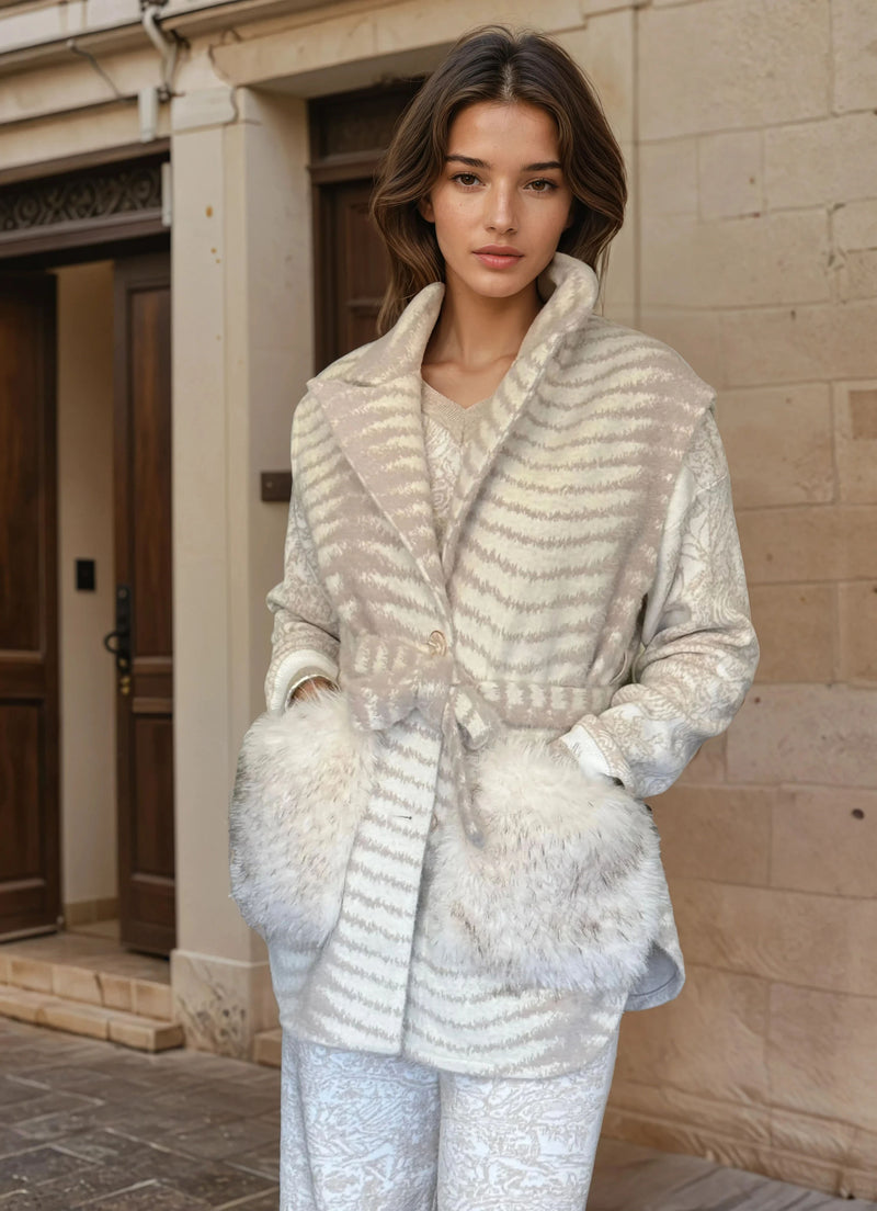 Stylish woman in neutral-toned fur-accented coat with waist tie and textured ivory pants, leaning against a stone wall.