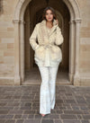 Fashion model wearing a chic cream fur-pocket jacket and white brocade pants, walking through an arched stone corridor.