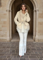 Fashion model wearing a chic cream fur-pocket jacket and white brocade pants, walking through an arched stone corridor.