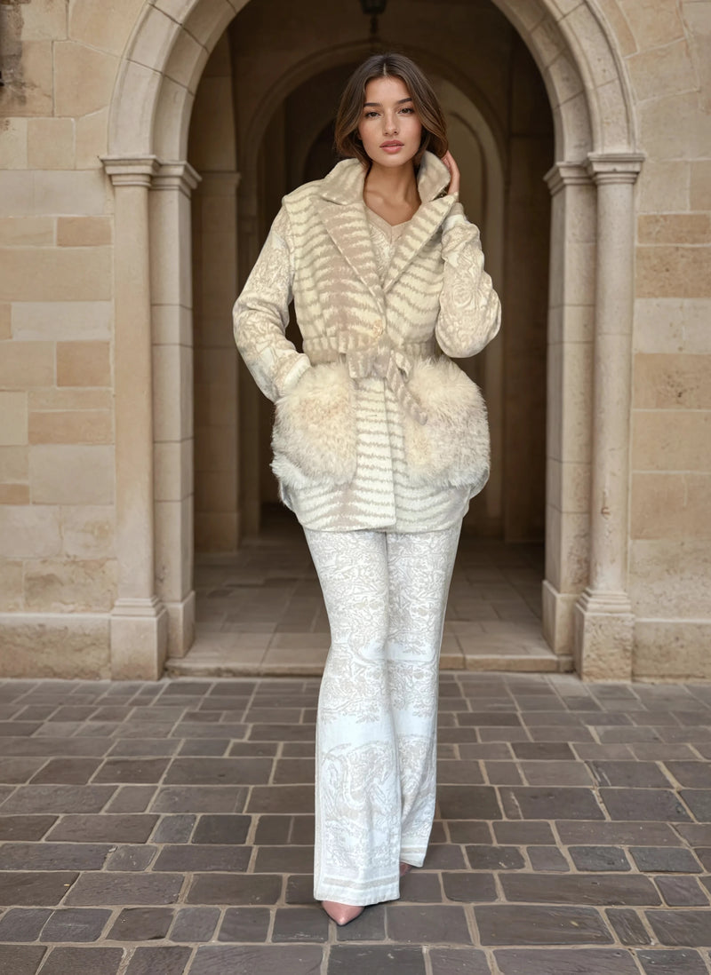 Fashion model wearing a chic cream fur-pocket jacket and white brocade pants, walking through an arched stone corridor.