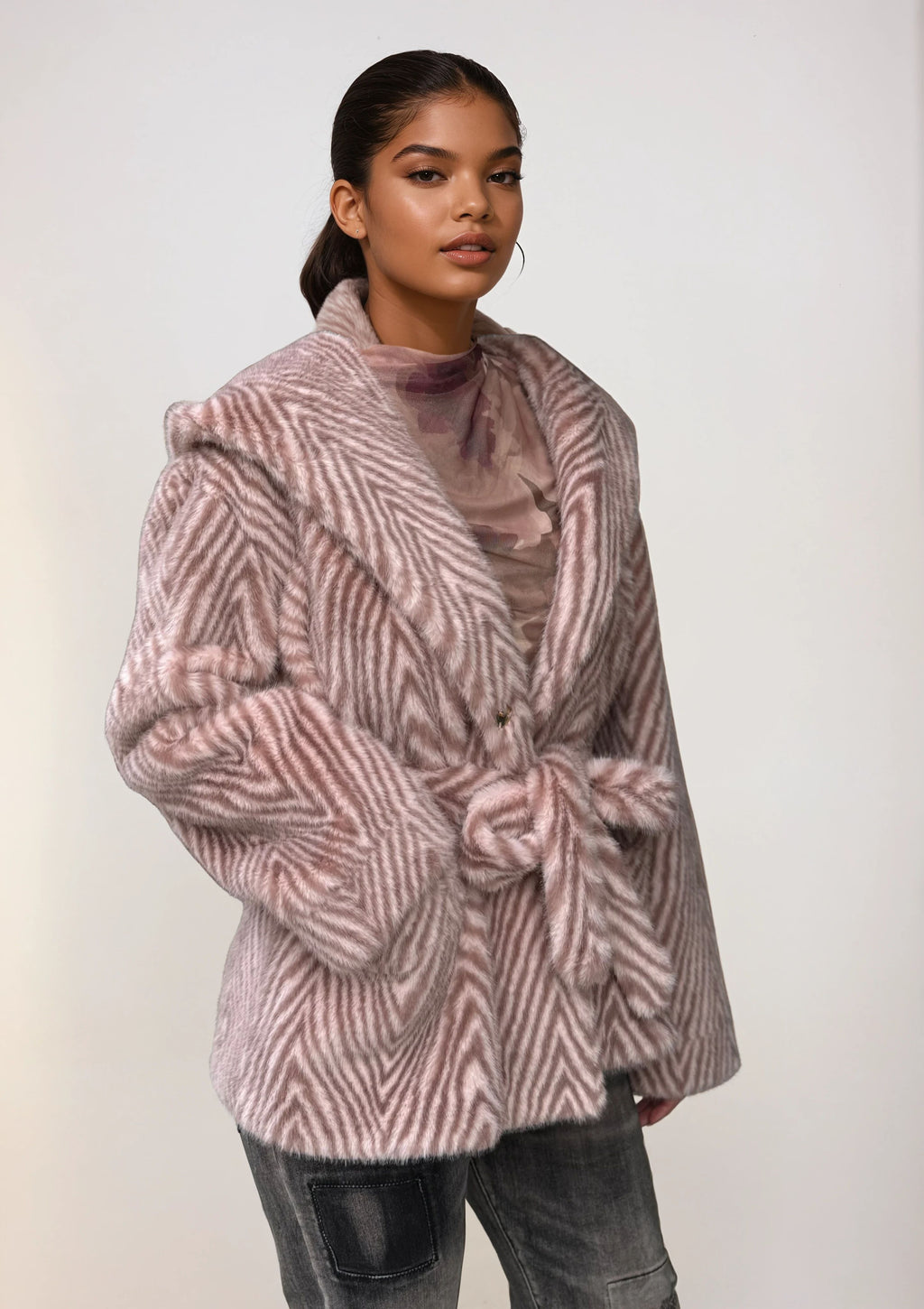 Model wearing a soft pink faux fur coat with chevron pattern and waist tie, paired with distressed gray jeans and a sheer top.