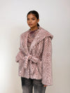 Stylish woman in a blush pink chevron faux fur jacket tied at the waist, layered over a mesh floral blouse and gray denim.