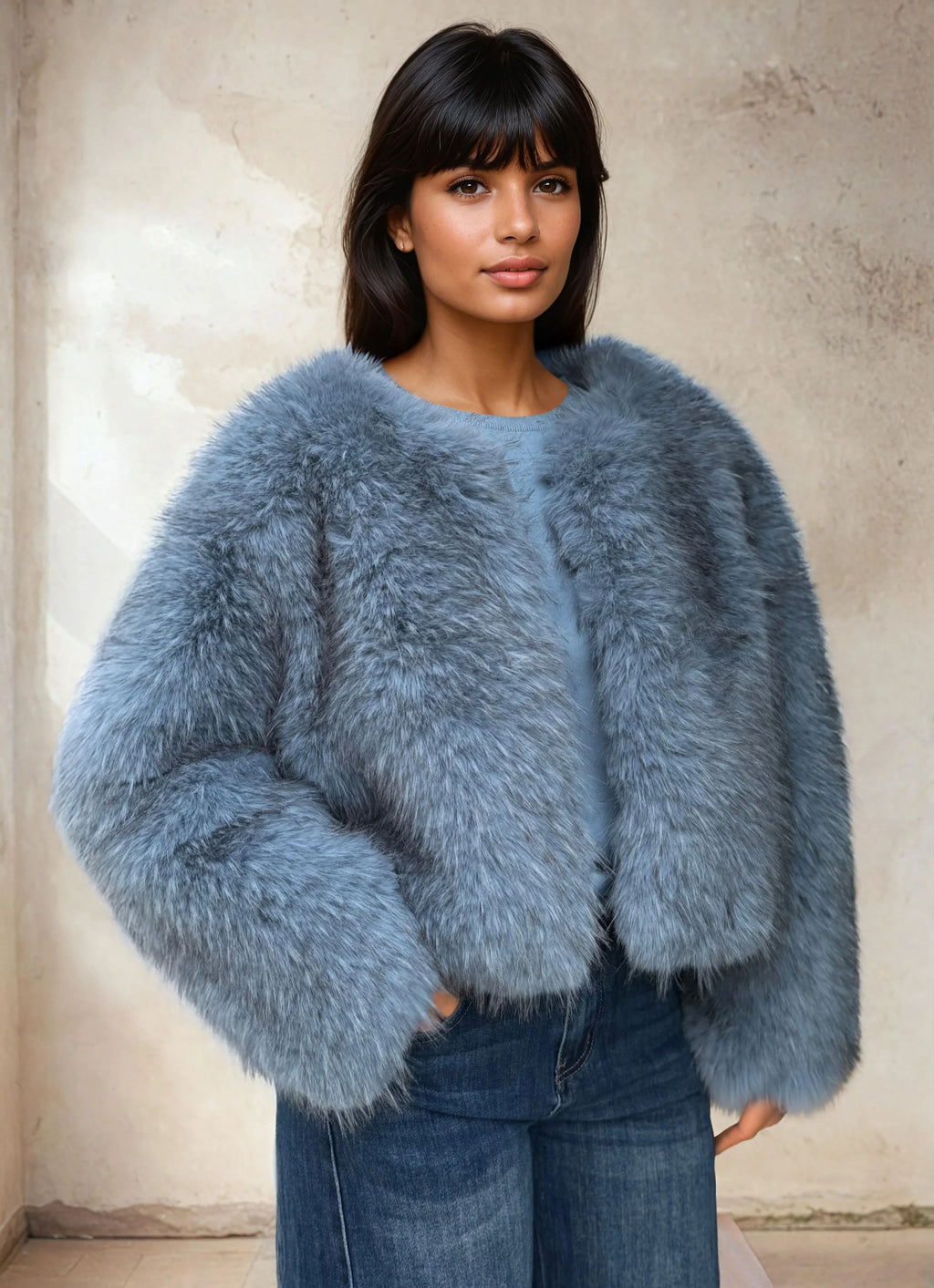 Woman wearing a cropped blue faux fur jacket styled with blue denim jeans in a minimalist indoor setting.