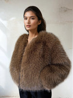 Woman wearing a luxurious brown faux fur jacket with long sleeves, posing against a neutral textured wall. She has natural makeup and sleek dark hair, styled simply for an elegant winter fashion look.