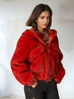 Model wearing a bold blood orange faux fur hooded jacket with oversized sleeves and front pockets, styled with black jeans in a neutral interior setting.