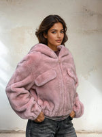 Close-up of model in a soft pink faux fur jacket with front pockets and hood, styled with grey jeans, standing against a textured beige wall.