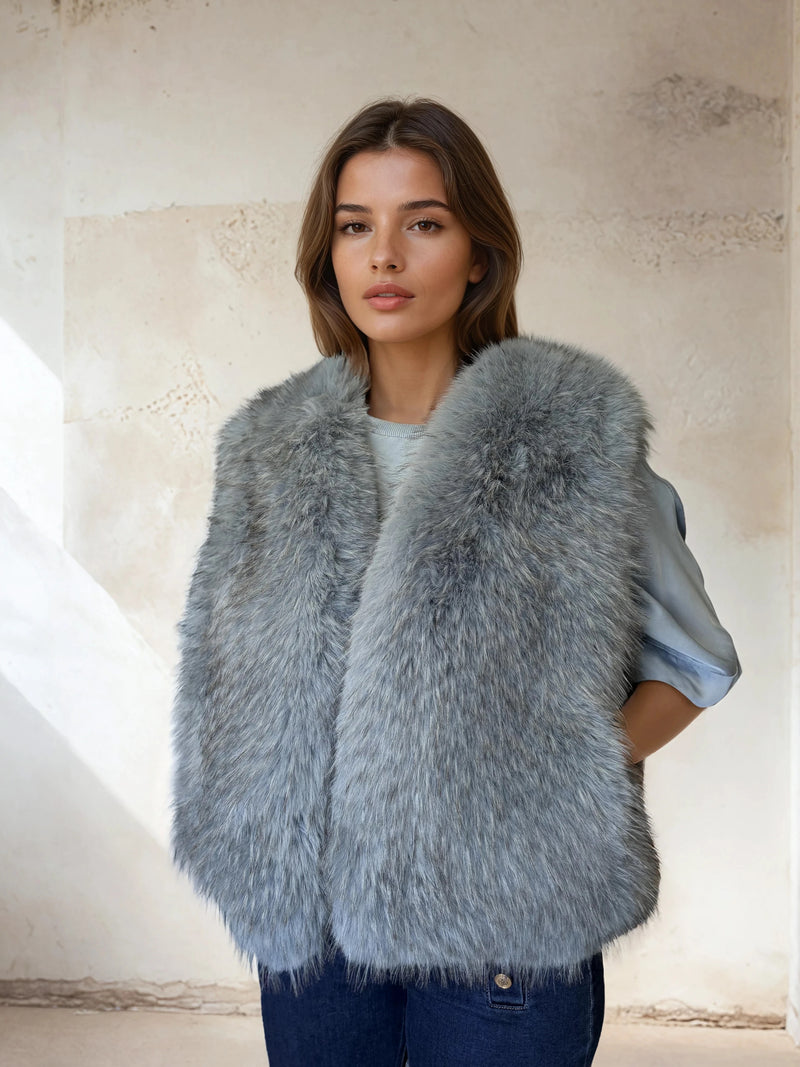 Woman dressed in a cool gray-blue fur vest over a light blue blouse and dark jeans, posing against a warm neutral textured wall with soft natural lighting.