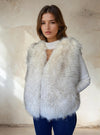 Woman modeling an elegant white fur vest with subtle gray shading, worn over a white knit top with a cut-out neckline, paired with dark jeans.