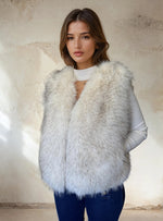 Woman modeling an elegant white fur vest with subtle gray shading, worn over a white knit top with a cut-out neckline, paired with dark jeans.