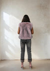 Back view of a woman in a light pink faux fur vest, gray jeans, and pink heeled boots, standing against a minimalist beige wall with natural light.