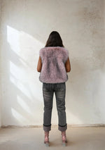 Back view of a woman in a light pink faux fur vest, gray jeans, and pink heeled boots, standing against a minimalist beige wall with natural light.