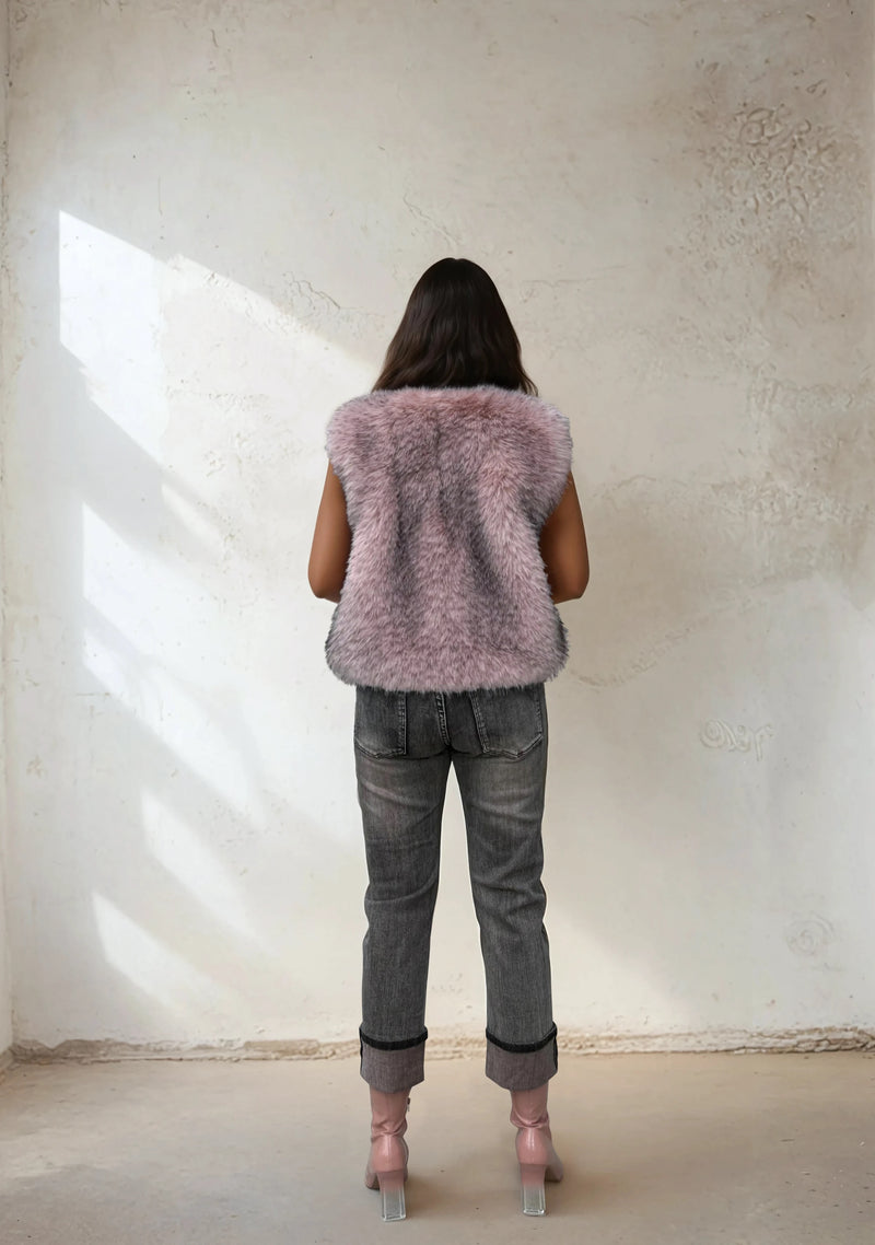 Back view of a woman in a light pink faux fur vest, gray jeans, and pink heeled boots, standing against a minimalist beige wall with natural light.