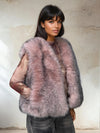 Woman wearing a soft blush pink fur vest with gray undertones, styled over a sheer patterned top and gray jeans, posing in front of a textured beige wall.