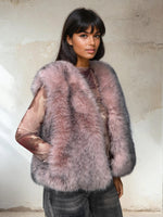 Woman wearing a soft blush pink fur vest with gray undertones, styled over a sheer patterned top and gray jeans, posing in front of a textured beige wall.