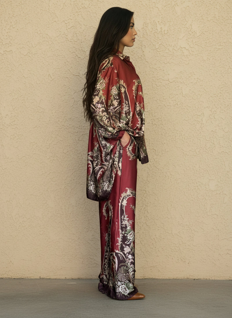 Side view of a woman in a burgundy silk two-piece set with ornate floral baroque patterns, wide-leg trousers, and oversized blouse. Elegant and comfortable fashion outfit.