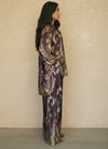Model wearing a luxurious silk set with a deep brown and olive baroque floral print, featuring a relaxed-fit shirt and wide-leg pants. Elegant resort wear outfit with a sophisticated pattern.