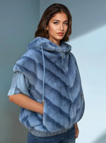 Woman wearing a light blue sleeveless faux fur hoodie with diagonal chevron texture, styled over a satin blouse and blue jeans, standing against a soft blue wall.