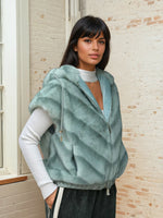 Model in a mint green sleeveless faux fur hoodie with drawstrings, styled over a white ribbed turtleneck and wide-leg trousers with side stripes.